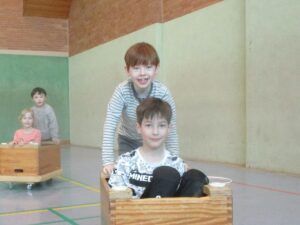 Zwei Kinder sitzen in hölzernen Kisten auf Rollen in einer Sporthalle.